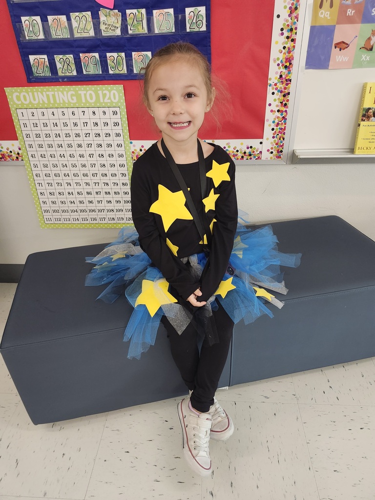 A young girl with blond hair wearing a black shirt with yellow stars and a blue tutu, smiling in a classroom setting with counting number charts and signs on the walls.