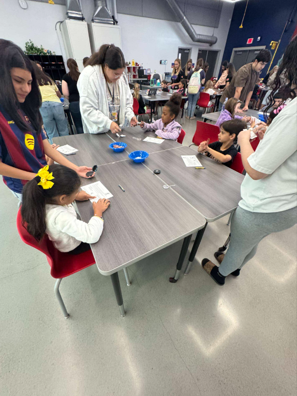 A group of people, likely students, working together at tables in a classroom or educational setting. Various art and craft supplies are visible on the tables.