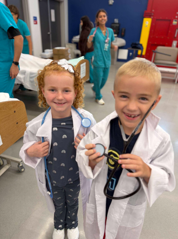 Two young children in medical attire, a girl with curly blonde hair and a boy with short blonde hair, both smiling and holding medical equipment.