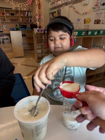 A young child sitting at a table, focused on pouring a red substance into a container of milkshake or smoothie.