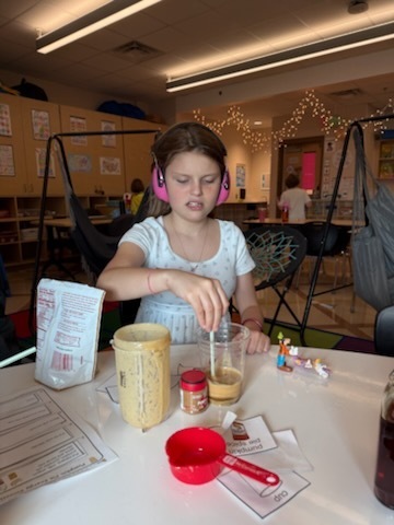 A young girl with headphones sits at a table with various cooking and baking supplies, including a measuring cup, mixing bowl, and spoon.