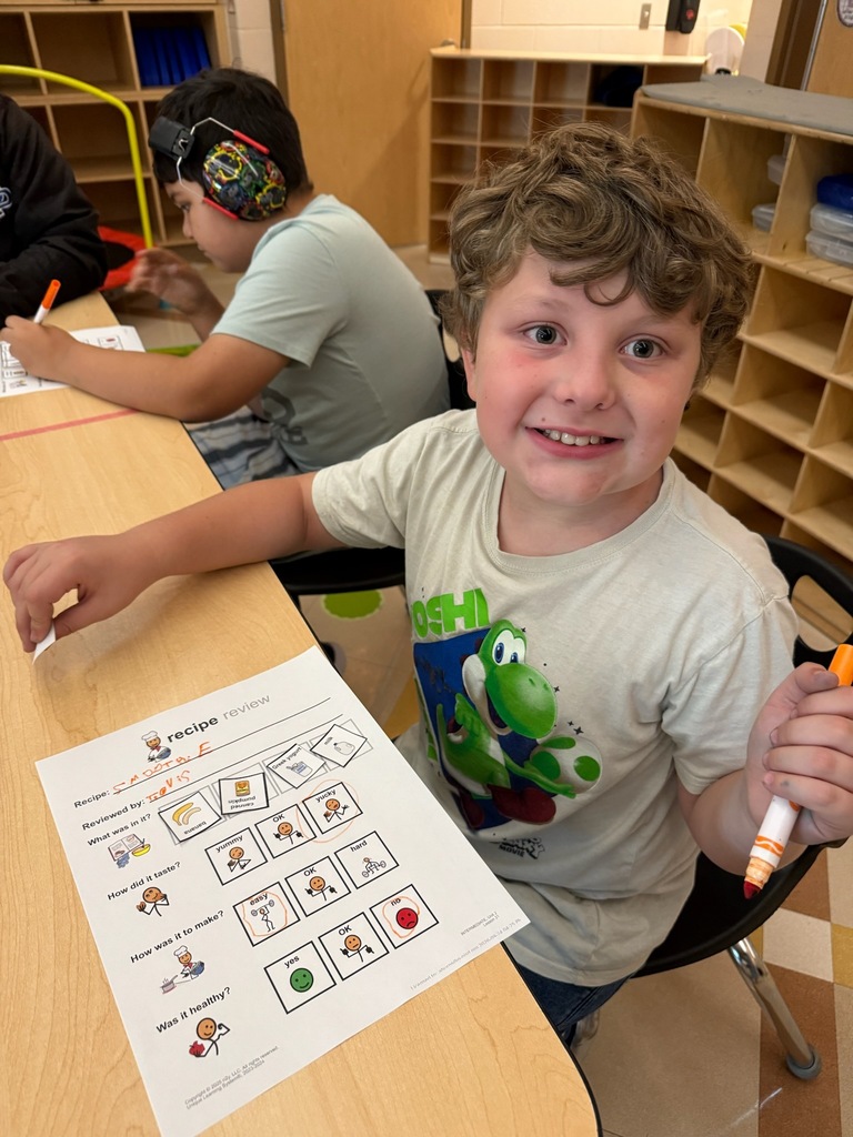 A young boy with curly hair smiling while seated at a table and working on a classroom worksheet.