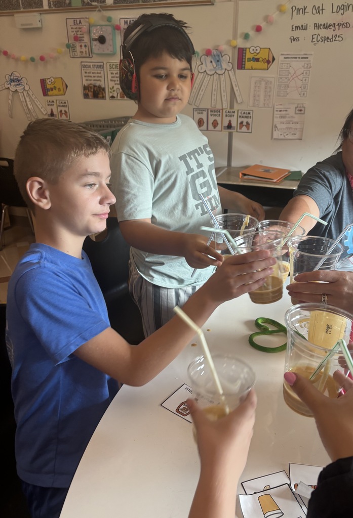 Three children in a classroom, two boys and a girl, sitting at a table and mixing drinks in clear cups.