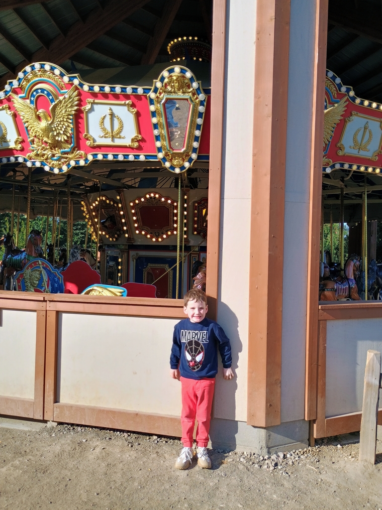 Young child posing outside of a carousel on a sunny day. Child has red sweatpants and a blue sweatshirt, smiling for a photo.