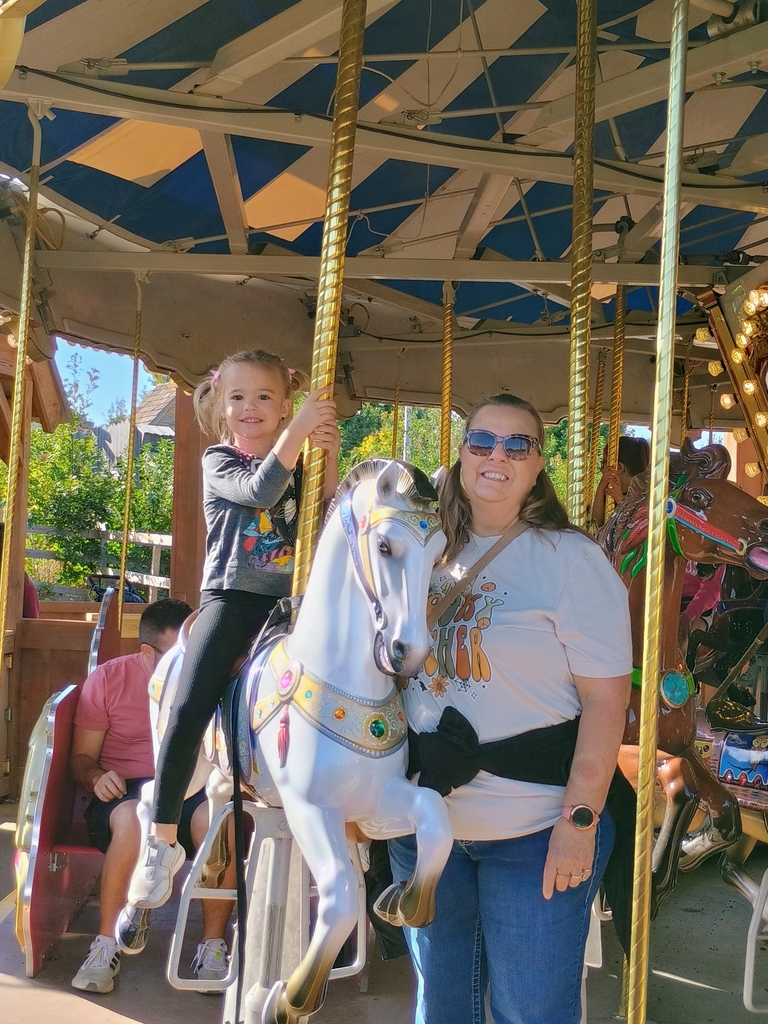 Young child and teacher posing on a carousel. Child is riding a white horse on the carosel. Subjects are smiling at the camera.
