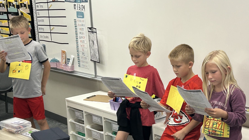 four students standing at the front of the classroom reading from papers like a readers theatre.