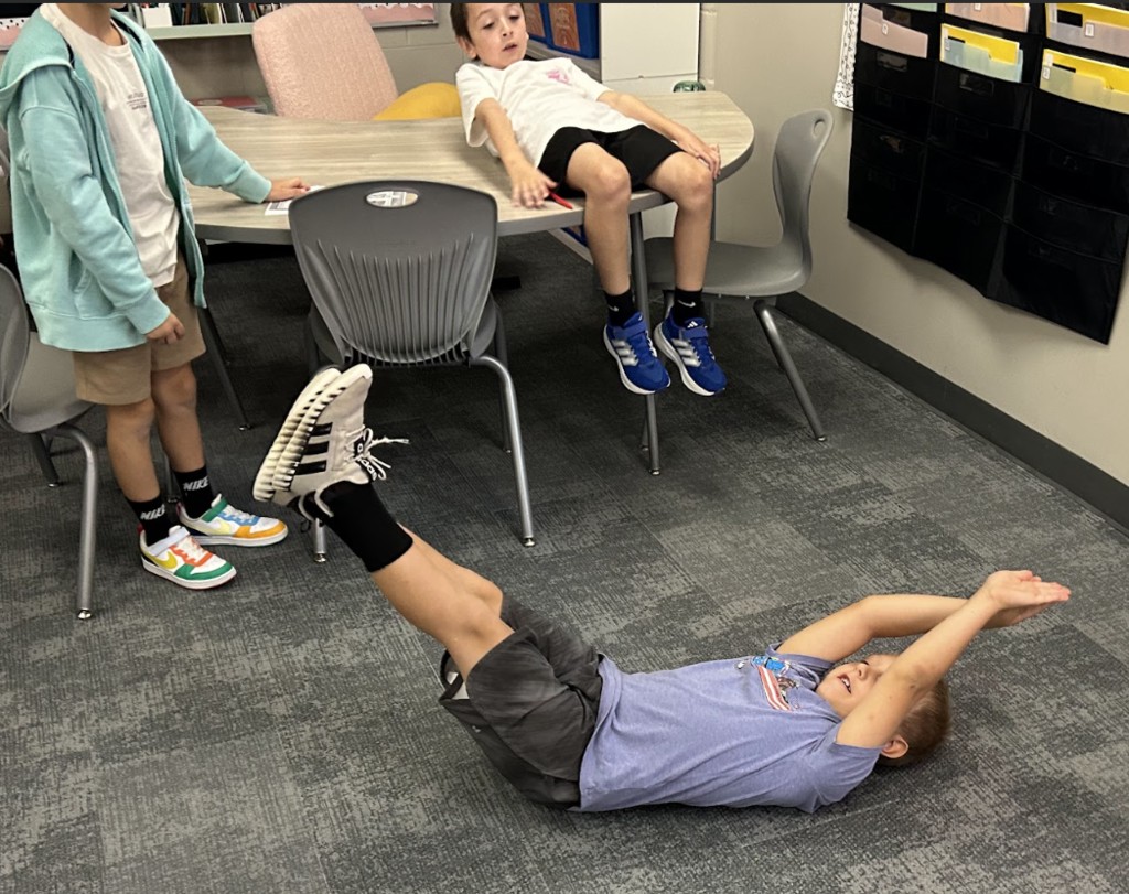 A student is acting out a scene in a book by laying on his back on the floor of a classroom.
