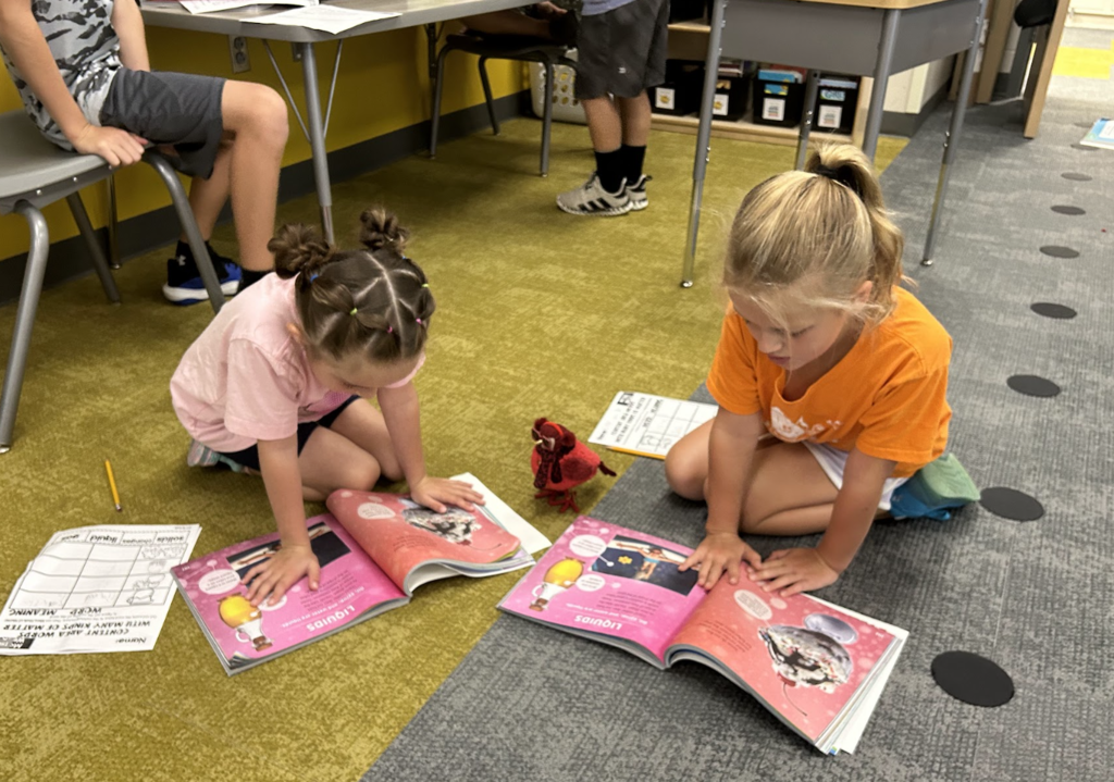 two female students sit on the floor in a classroom reading from their books together.