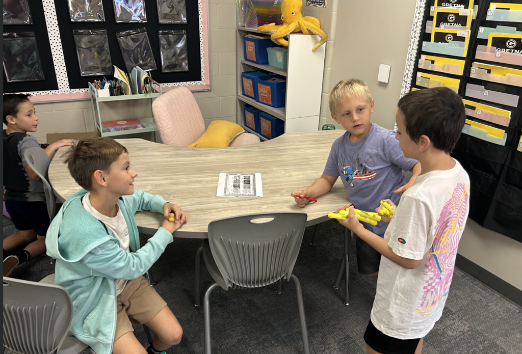 four students around a half circle table in a classroom. One student is holding out highlighters as thought he is going to pass them out to his friends.