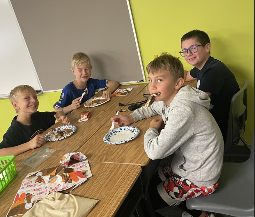 Photo of four boys eating pancakes. One boy has the entire pancake in his mouth and the other three are laughing.