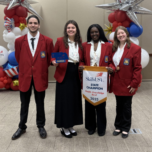 A group of 4 Gretna East students in red blazers pose with the Skills USA State Champion banner for winning the Health Knowledge Bowl