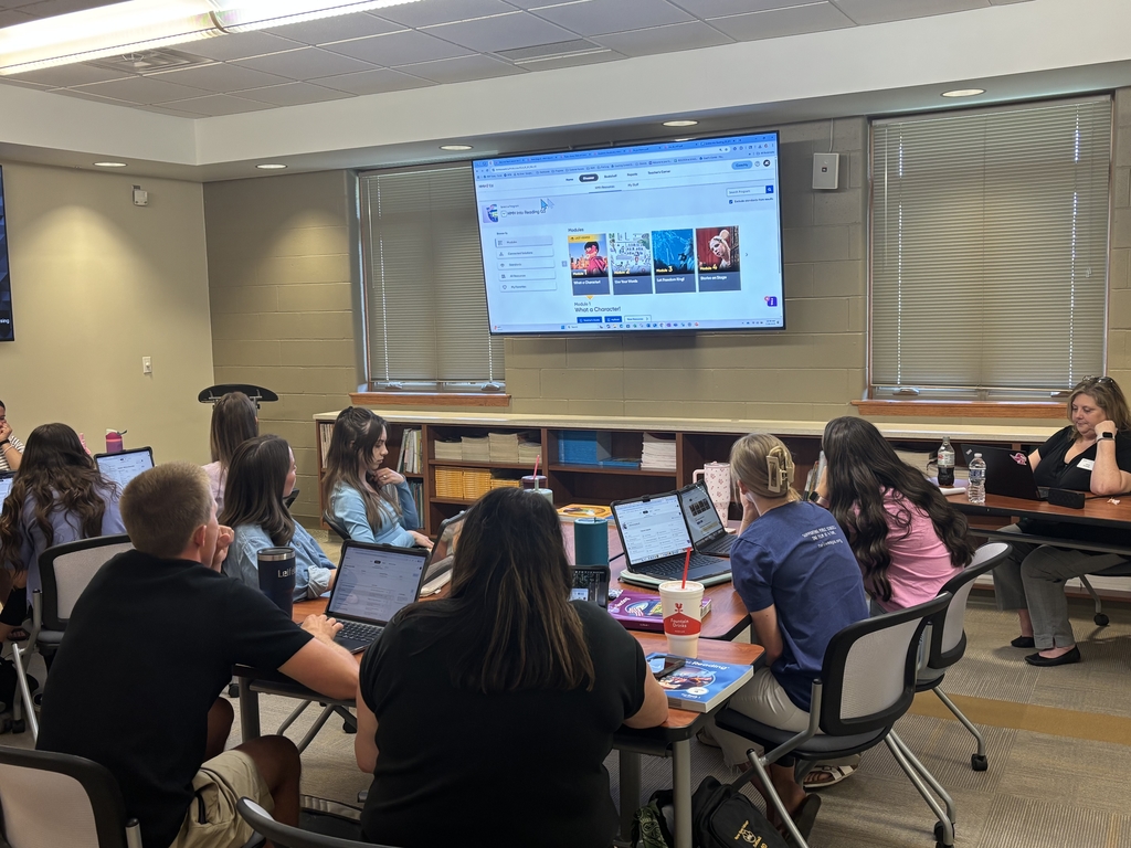 New staff engaged in learning about curriculum around tables with laptops and tv displaying reading choices