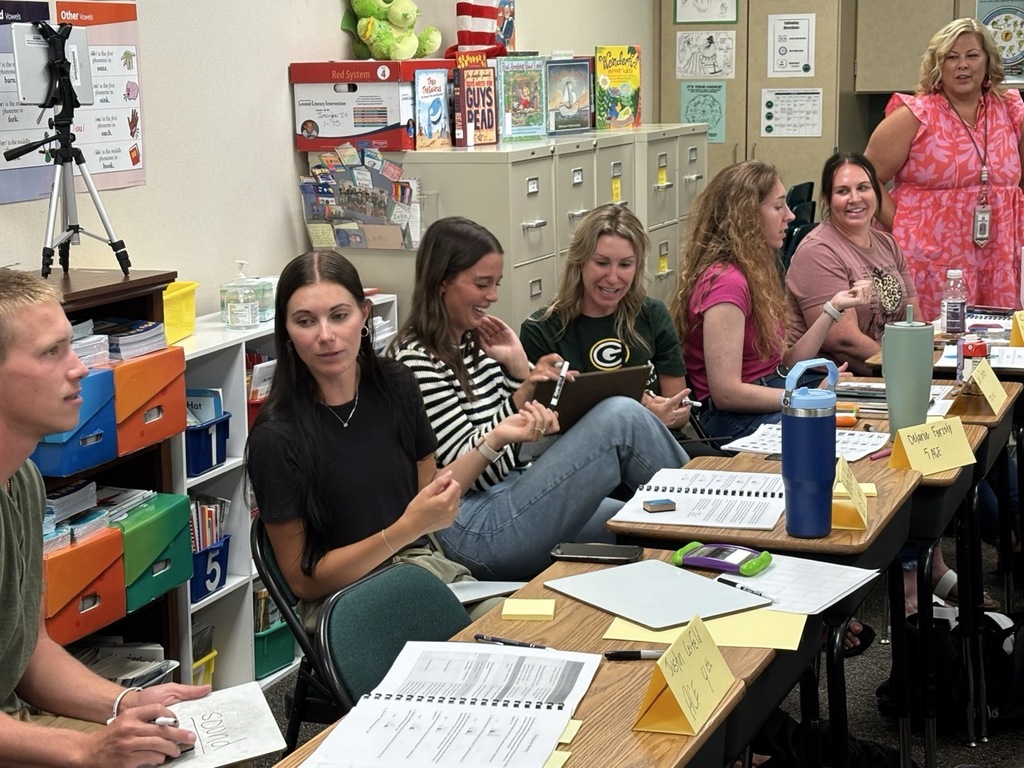 group of seven teachers sitting in classroom at desks collaborating and smiling with each other