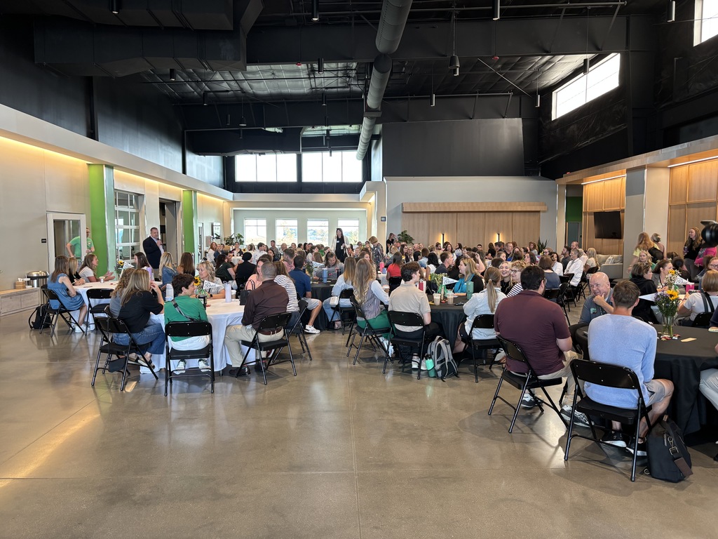 A large group of new staff members seated around tables in a large room while Superintendent Travis LIghtle presents in front