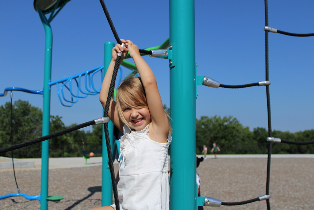 A smiling young girl climbs on rope play equipment on the sunny playground during recess time.