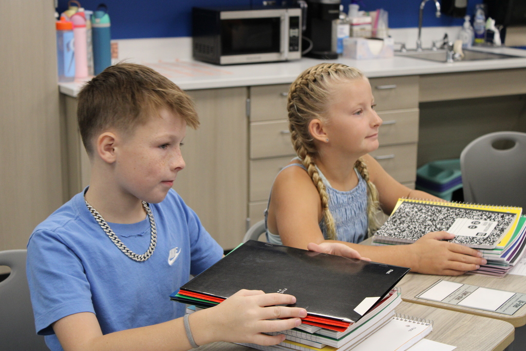 Two kids in class look ready to conquer the school year with towering stacks of notebooks.