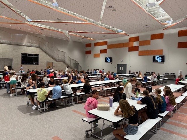 About 30 6th graders eating lunch in the cafeteria with stairs in the background