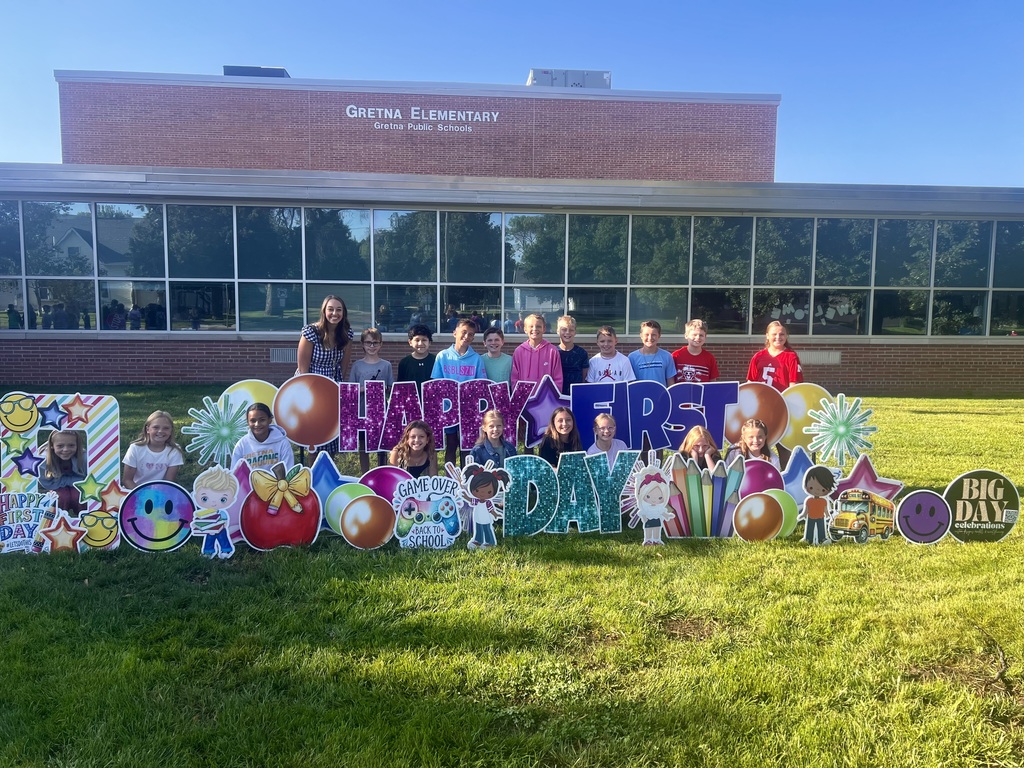 Group of students standing in front of a brick school building with "Happy First Day" cards pushed into the grass.