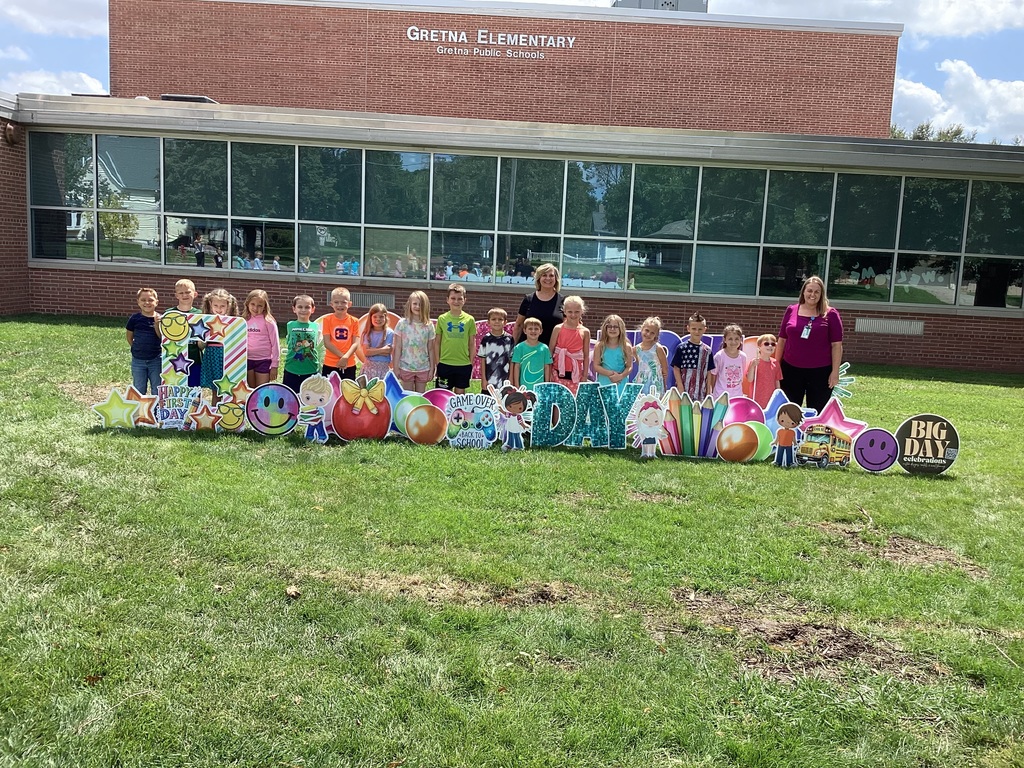 Group of students standing in front of a brick school building with "Happy First Day" cards pushed into the grass.
