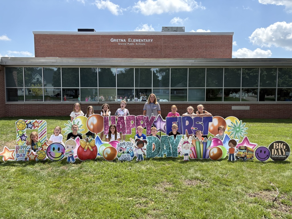 Group of students standing in front of a brick school building with "Happy First Day" cards pushed into the grass.