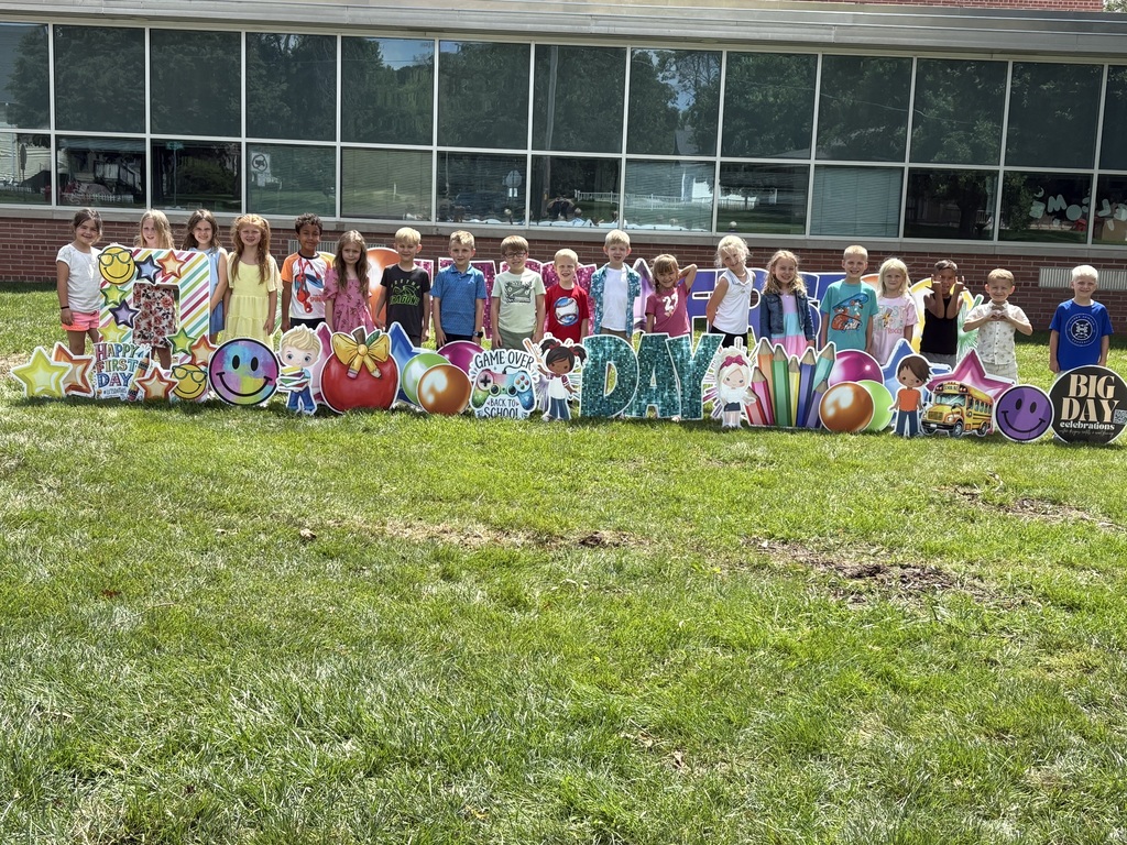 Group of students standing in front of a brick school building with "Happy First Day" cards pushed into the grass.