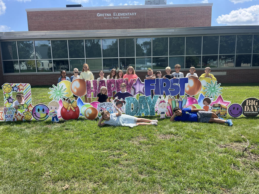 Group of students standing in front of a brick school building with "Happy First Day" cards pushed into the grass.