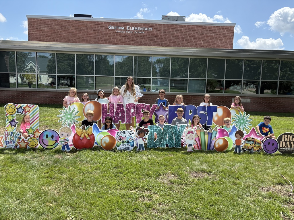 Group of students standing in front of a brick school building with "Happy First Day" cards pushed into the grass.