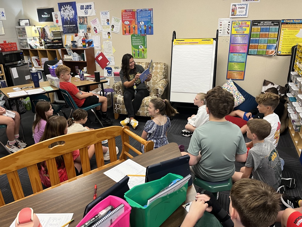 A group of elementary students sitting and listening to a book that is read by a woman.