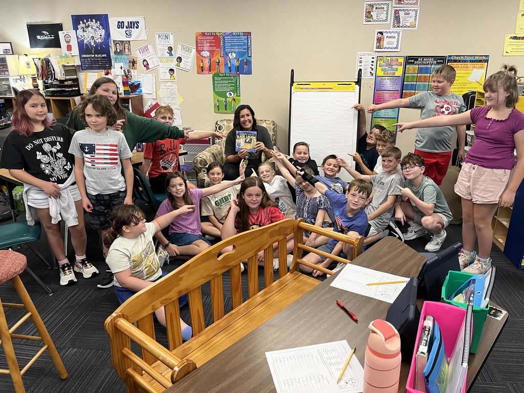 Group of students, some standing and some sitting, pointing at an adult holding a book.