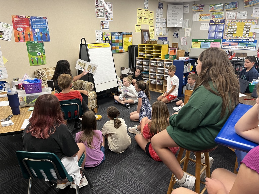 A group of elementary students sitting and listening to a book that is read by a woman.