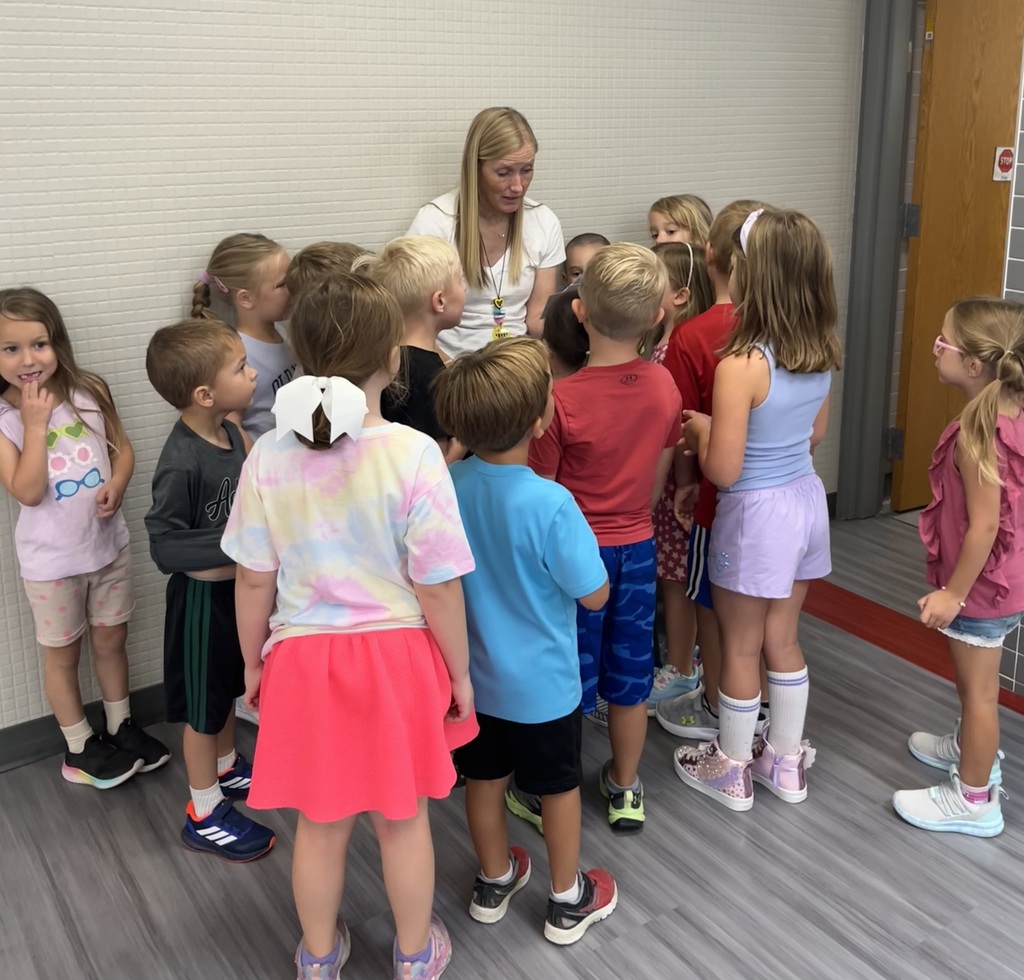 A group of children gathered around a woman in a hallway, listening intently.