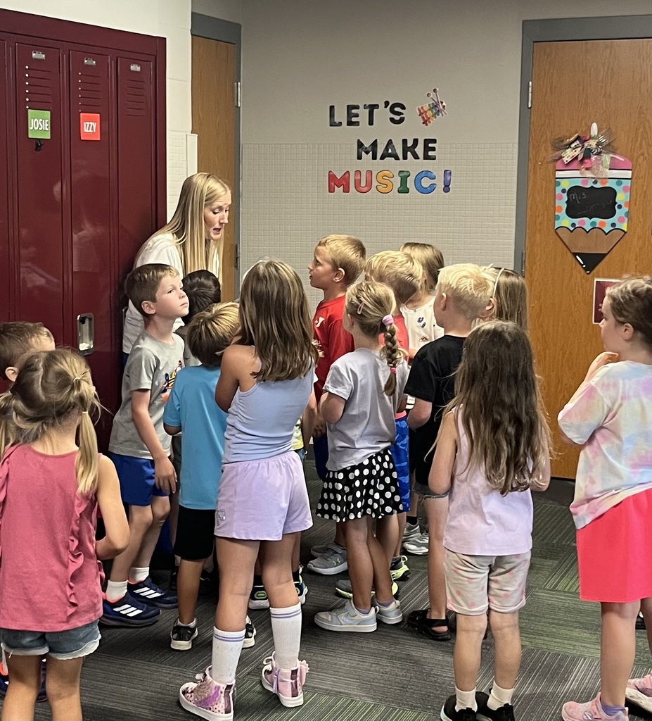A group of children gathered around a woman in a hallway, listening intently.