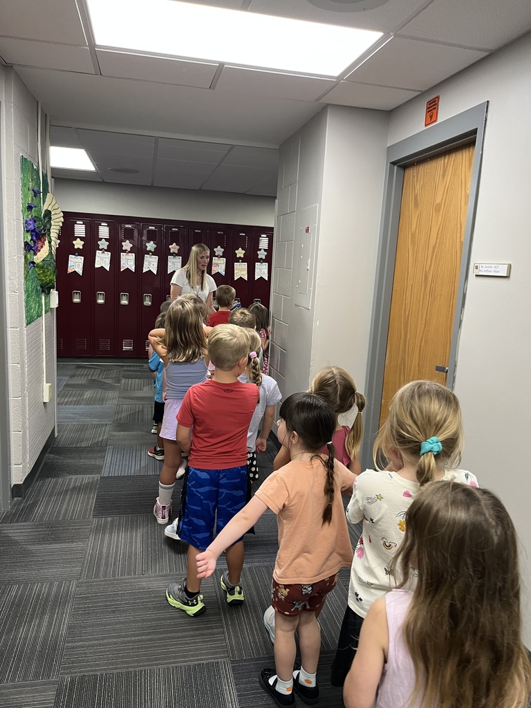 A group of children gathered around a woman in a hallway, listening intently.