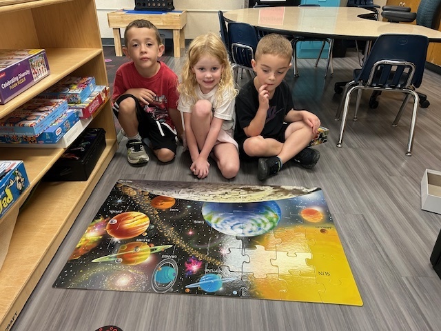 Three kids sit on tile in front of a puzzle of the solar system.