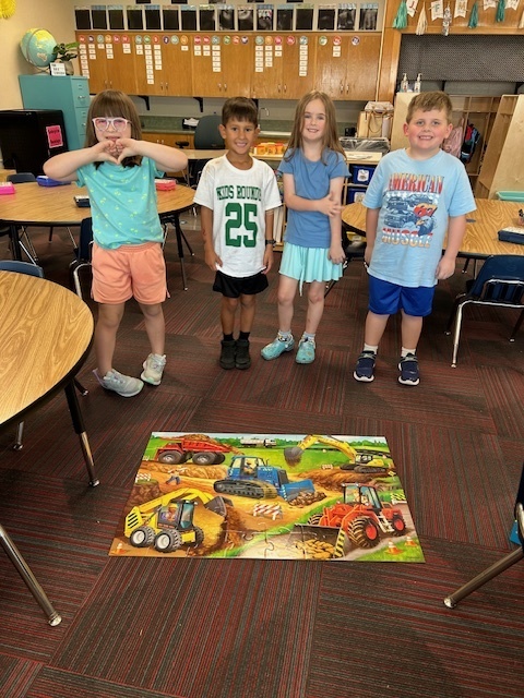 Four kids stand on a rug in front of a puzzle of heavy machinery.