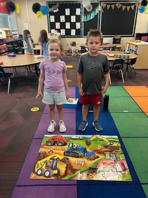 A boy and girl standing on a colored rug in front of a puzzle of heavy machinery.