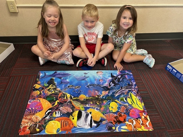 Three kids sit on a rug in front of a puzzle of fish in the water.
