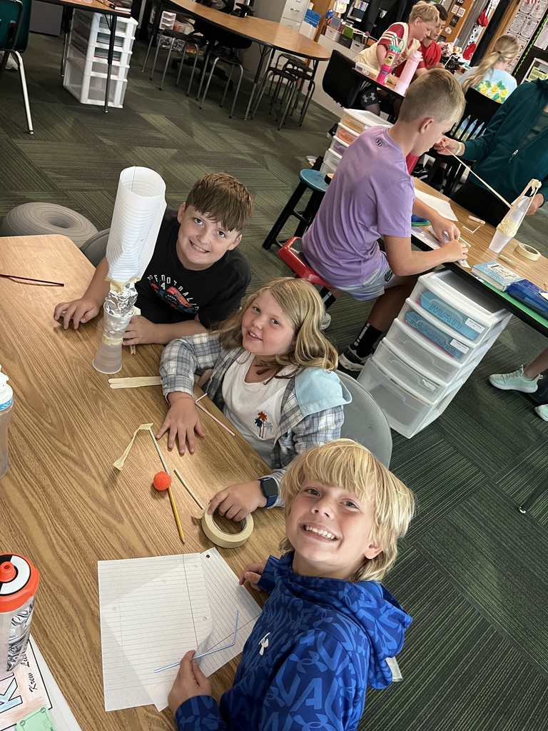 Three kids sit at a table smiling while holding various supplies like tape and straws.