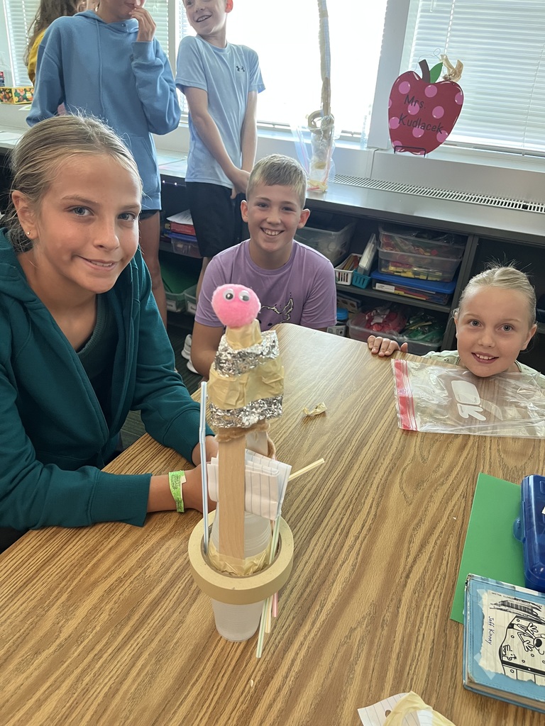 Three kids sit at a table smiling while holding various supplies like tape and straws in front of a contraption made out of bottles and tape.