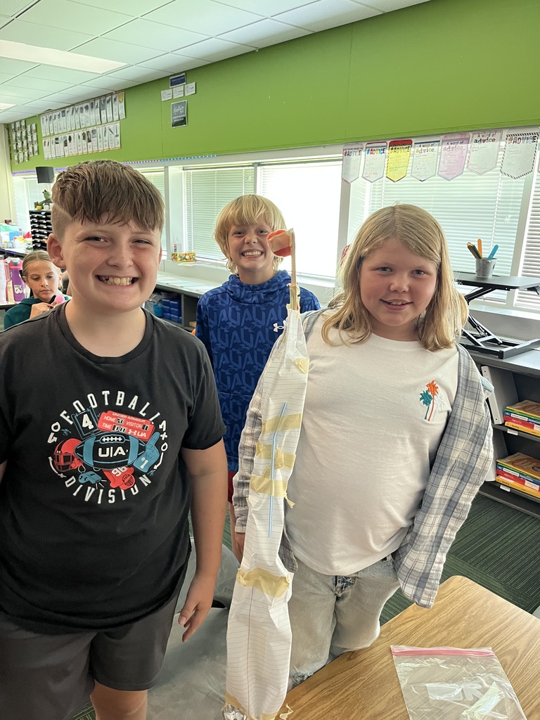 Three kids stand at a table smiling while standing in front of a tall tower made out of tape and a water bottle.