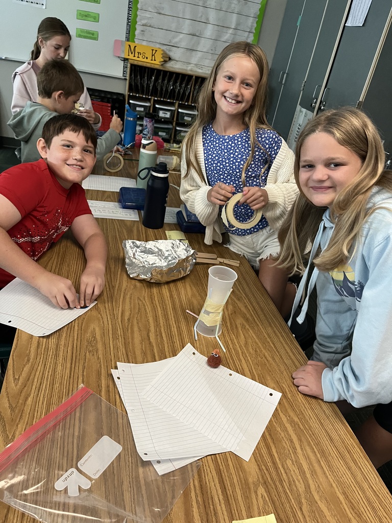 Three kids sit at a table smiling while holding various supplies like tape and straws