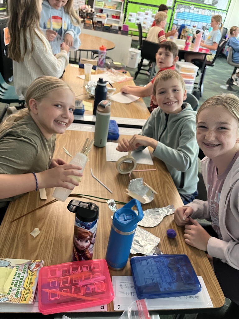 Four kids sit at a table smiling while holding various supplies like tape and straws