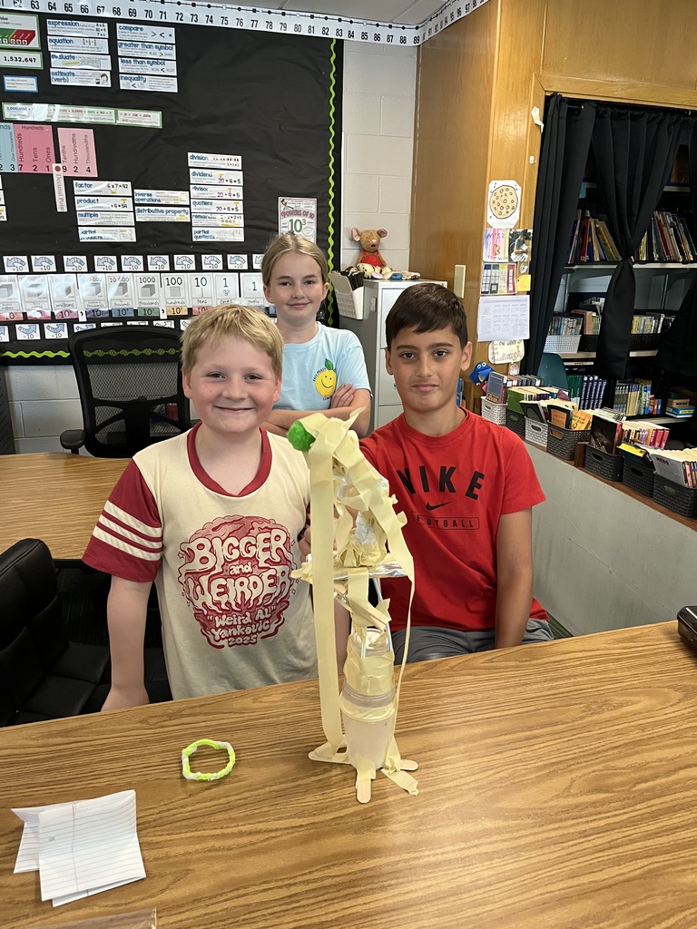 Three kids stand at a table smiling while standing in front of a tall tower made out of tape and a water bottle.