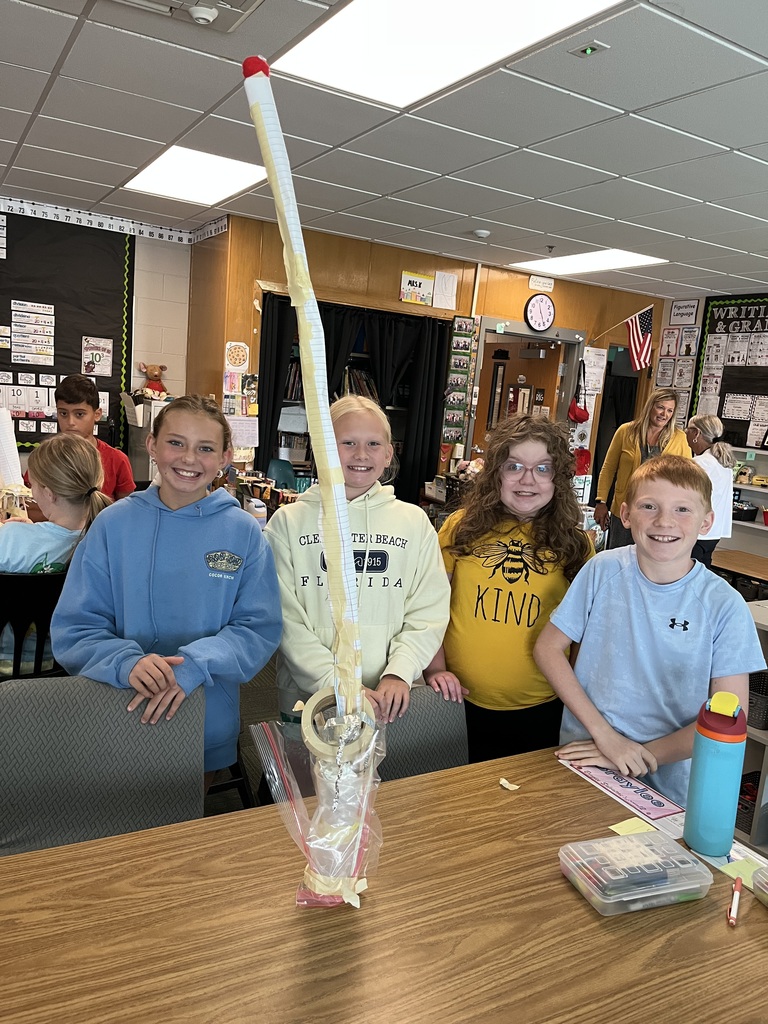 Four kids stand at a table smiling while standing in front of a tall tower made out of tape and a water bottle.
