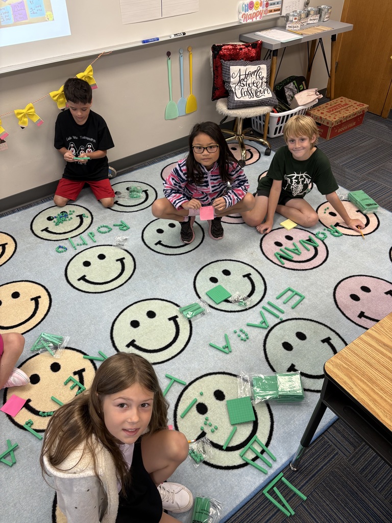 Four students sit on a rug that has green place value blocks with their names spelled out.