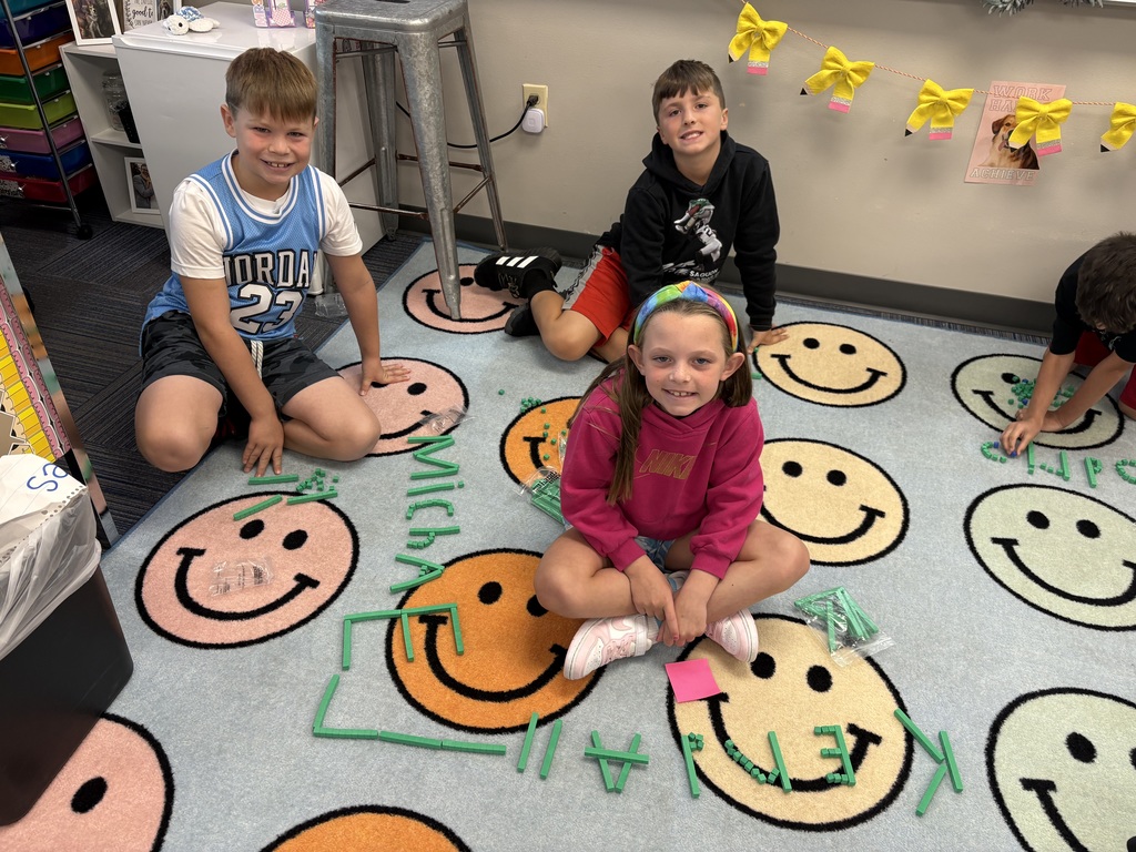 Three students sit on a rug that has green place value blocks with their names spelled out.