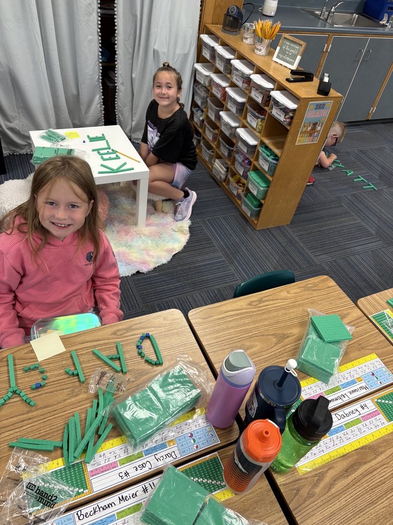 Two students sit at desks that have green place value blocks with their names spelled out.