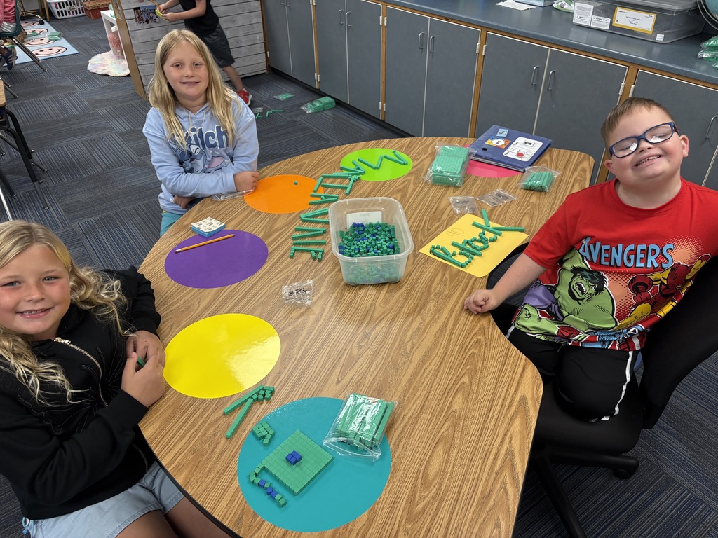Three students sit at a table that has green place value blocks with their names spelled out.