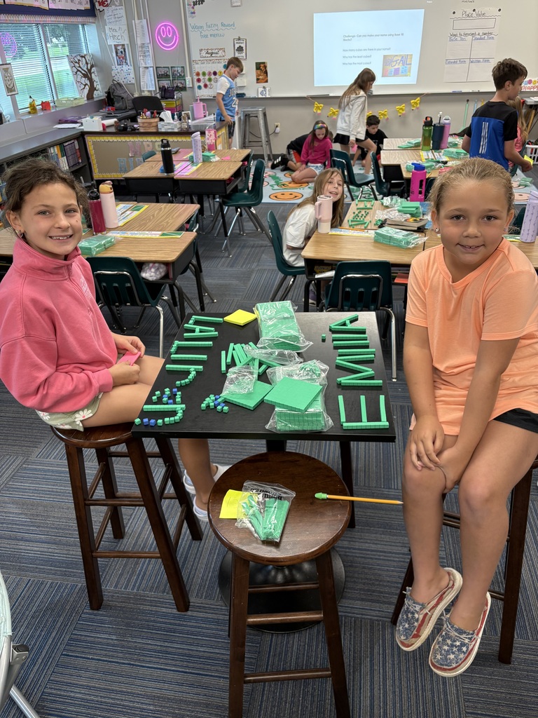 Two students sit at desks that have green place value blocks with their names spelled out while other students look on in the background.