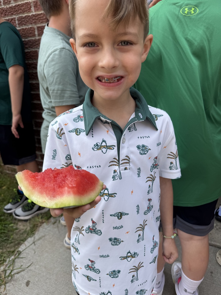 Boy holds up a chunk of watermelon while smiling at the camera while getting his photo taken.